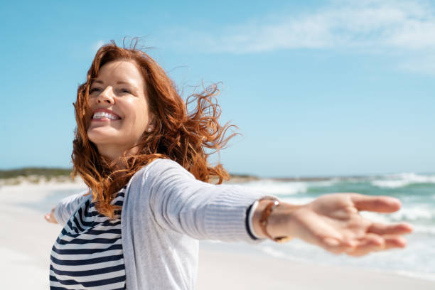 Mature woman enjoy breeze at beach Happy mature woman with arms outstretched feeling the breeze at beach. Beautiful middle aged woman with red hair and arms up dancing on beach in summer during holiday. Mid lady in casual feeling good and enjoying freedom with open hands at sea, copy space. arms outstretched stock pictures, royalty-free photos & images