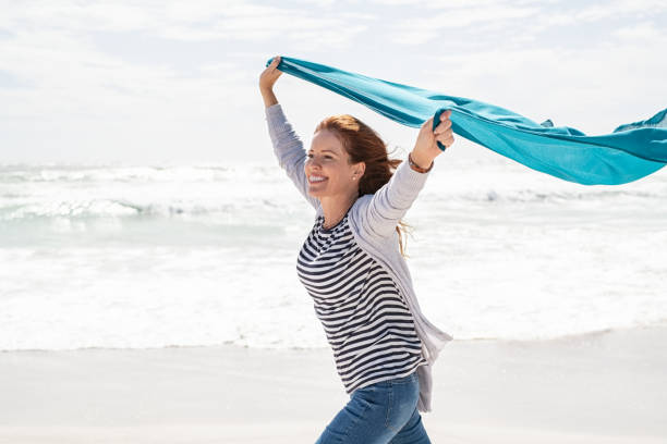 mujer madura corriendo en la playa con bufanda azul - jumping women running vitality fotografías e imágenes de stock