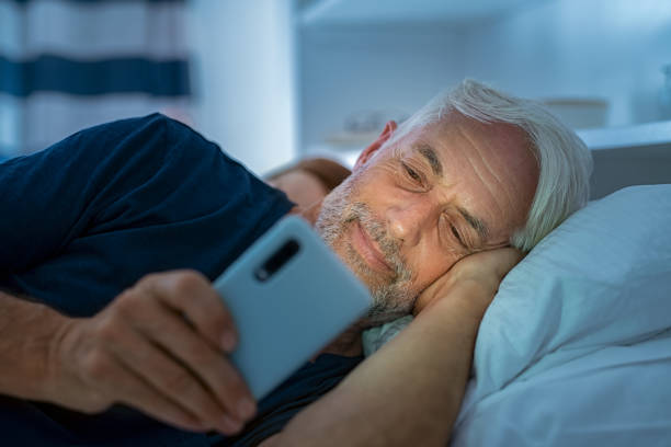 Mature man texting on mobile phone late at night Senior man watching video on smart phone while in bed. Smiling mature man lying on bed using cellphone in the dark at home. Addict man awake using phone for chatting late in night. old man pajamas photos stock pictures, royalty-free photos & images