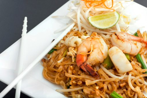 Seafood pad Thai dish of fried rice noodles on a square white plate with chopsticks and grated carrot garnish.
