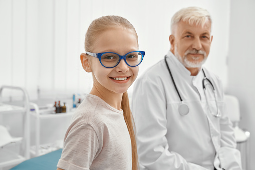 Selective focus of girl in eyeglasses looking at camera and smiling, eldery male doctor with stethoscope on neck on background. Little patient and therapist sitting on couch in modern hospital office.