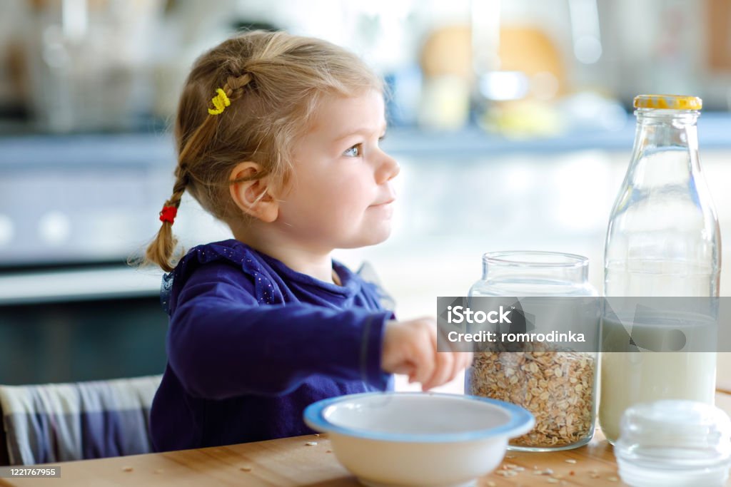 Entzückende Kleinkind Mädchen essen gesunde Hafermahlzeiten mit Milch zum Frühstück. Nettes glückliches Baby Kind in bunten Kleidern sitzen in der Küche und Spaß mit der Zubereitung von Hafer, Müsli. Drinnen zu Hause - Lizenzfrei Blondes Haar Stock-Foto
