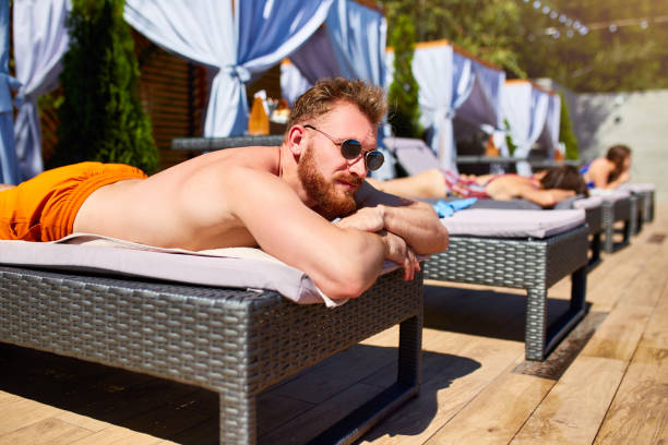 Man in sunglasses chilling on lounger with friends at poolside. Handsome guy relax at luxury resort on sunny day. People sunbathing on summer vacation at holiday villa Man in sunglasses chilling on lounger with friends at a poolside. Handsome guy relax at luxury resort on sunny day. People sunbathing on summer vacation at holiday villa. chaise longue woman stock pictures, royalty-free photos & images