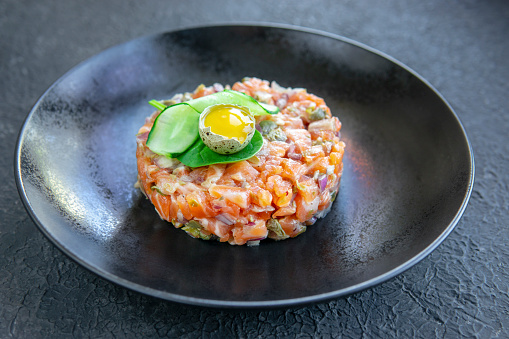 Fresh salmon tartar with quail egg, cucumber, greens on black table, textured dark table. Seafood raw concept. Copy space.