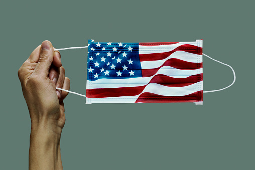 closeup of man holding a surgical mask patterned with the flag of the United States on a gray background