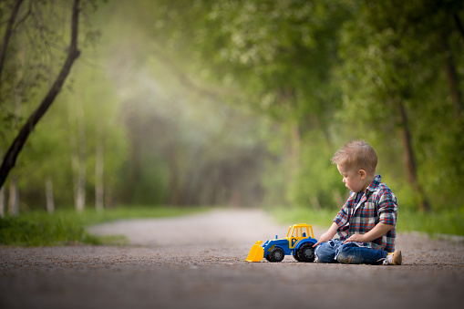 Portrait of cute kid boy sitting on the ground and playing with colorful tractor and sand in the park. Child playing outdoors. Lifestyle concept.