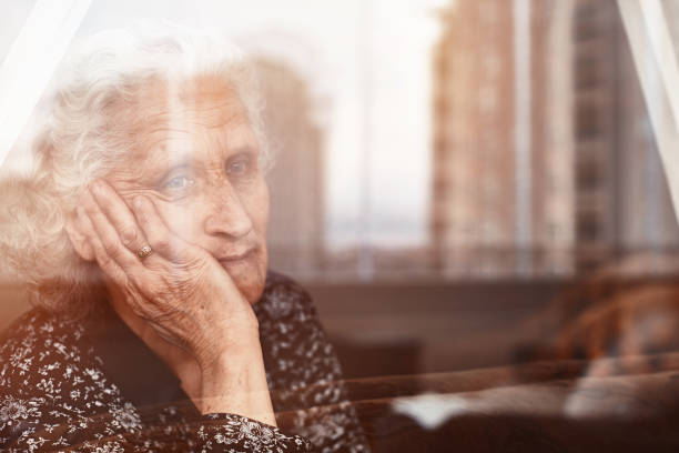 mujer anciana sentada sola y mirando tristemente fuera de la ventana - pensive senior adult looking through window indoors fotografías e imágenes de stock