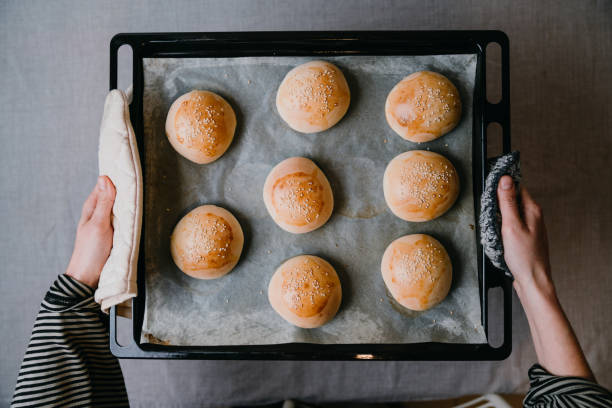 Young woman holding a baking tin of freshly cooked burger buns Young woman holding a baking tin of freshly cooked burger buns. High angle view. BREAD ROLLS stock pictures, royalty-free photos & images