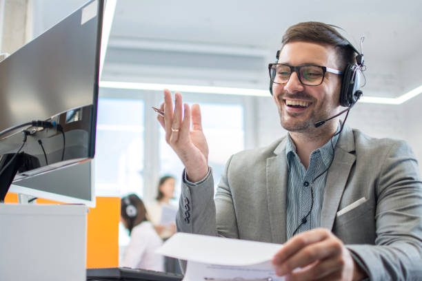 istock Smiling friendly handsome young male call centre operator wearing eyeglasses working on computer and checking paper documents at his workplace in the office. 1221800084