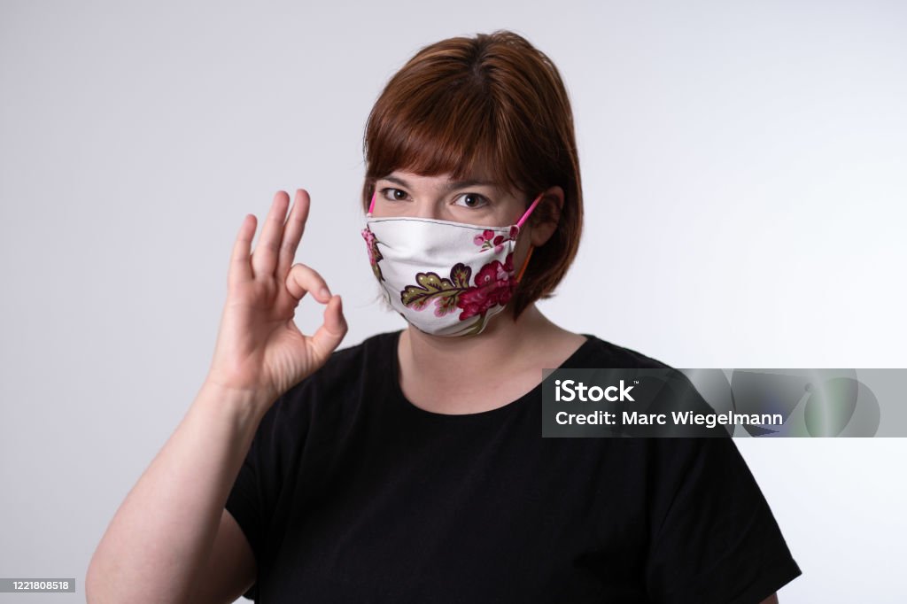 Young red haired woman putting on self made face mask with rose pattern Young woman is using a diy facemask with rose patterns in front of a white background to protect from the Coronavirus Covid-19. She is showing with a finger gesture that everything is fine. 30-39 Years Stock Photo