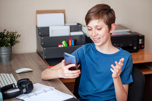 A teenage boy makes a video call using his mobile phone to his friends, classmates, and relatives. Joyfully shares the news at home in the room at the table.