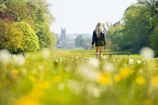 Pretty blond teenage girl walking through a bokeh of long grass and dandelions in Cirencester Park, Cirencester in the Cotswolds, UK