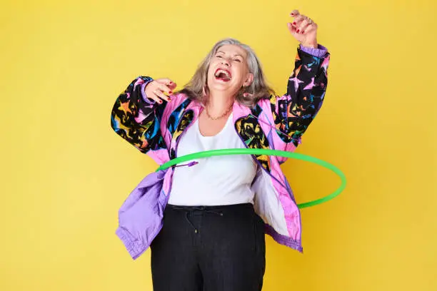 Cropped shot of a cheerful and stylish senior woman playing with a hula hoop against a yellow background