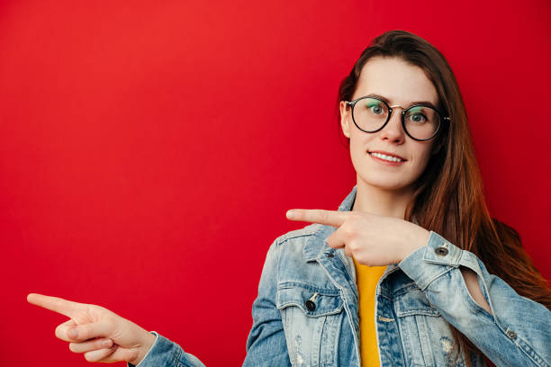istock Beautiful brunette woman in eyeglasses, points with hands aside, shows copy space for your advertisement or promotional text, dressed in denim jacket, isolated over red background. Promotion concept 1221823473