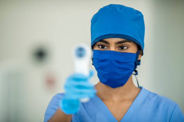 Middle Eastern doctor is in focus as she holds up a thermometer while wearing a mask
