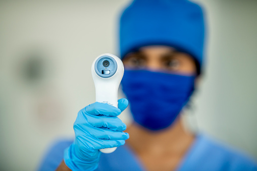 Middle Eastern doctor is out of focus as she holds up a thermometer in focus while wearing a mask
