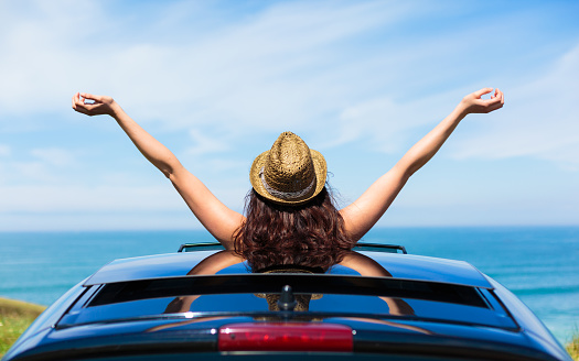 Rear view of relaxed woman on summer travel vacation to the coast  leaning out car sunroof towards the sea.
