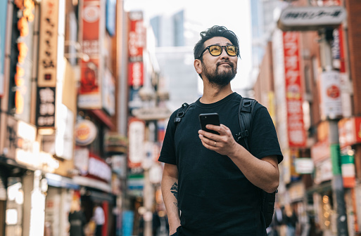 Asian man holding a smartphone and looking around in Tokyo famous neighborhood - Shibuya