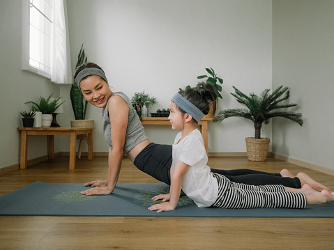 Asian little girl and her mother enjoying free time at home in the living room, practicing yoga together.