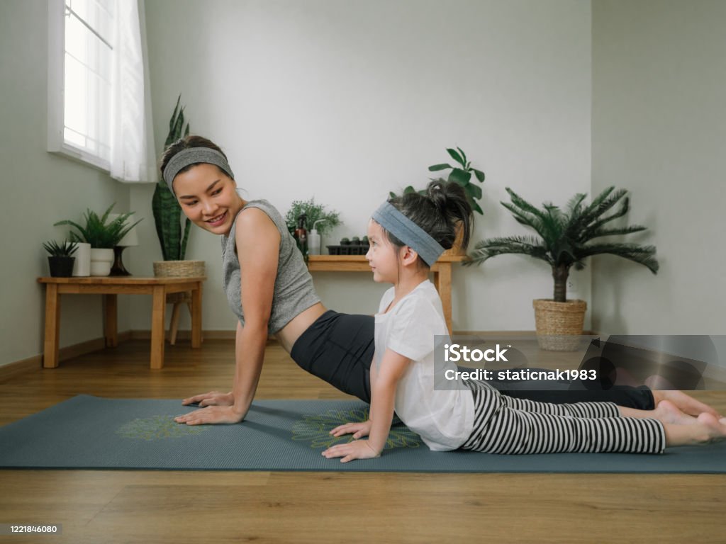 Mother doing on yoga mat with little daughter at home. Asian little girl and her mother enjoying free time at home in the living room, practicing yoga together. Child Stock Photo