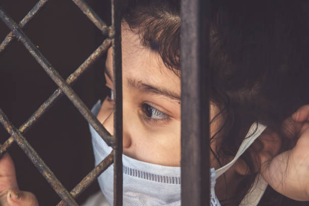 COVID-19 corona virus concept, little girl in medical mask looking through window at home. Sad kid during quarantine due to COVID. COVID-19 corona virus concept, little girl in medical mask looking through window at home. Sad kid during quarantine due to COVID curfew stock pictures, royalty-free photos & images