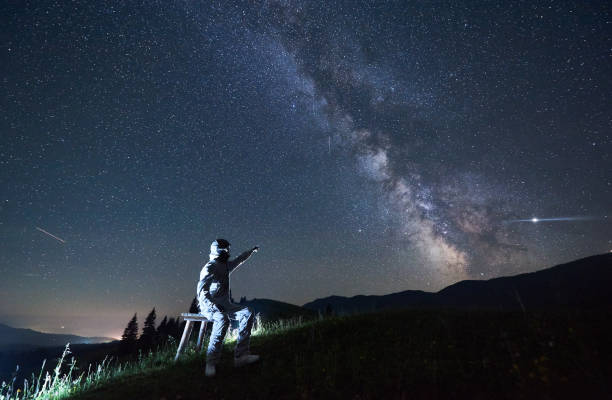 istock Illuminated man wearing white space suit and helmet sitting on a stool on a hill, pointing at starry sky 1221852168