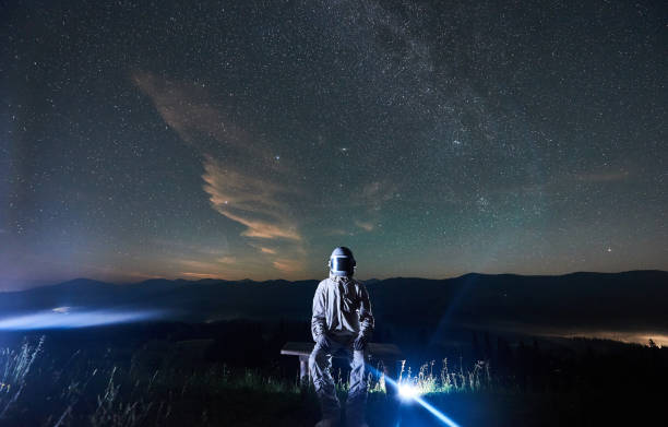 istock Illuminated man wearing white space suit and helmet sitting on a bench on a hill at night 1221852177