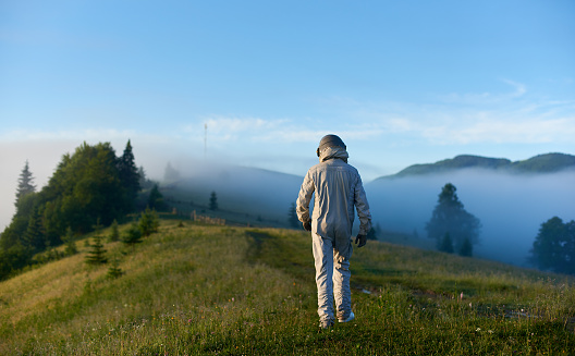 Back view of cosmonaut space traveler in space suit walking on green grass and heading to foggy hills with misty mountains and blue sky on background. Concept of astronautics, exploration and nature.