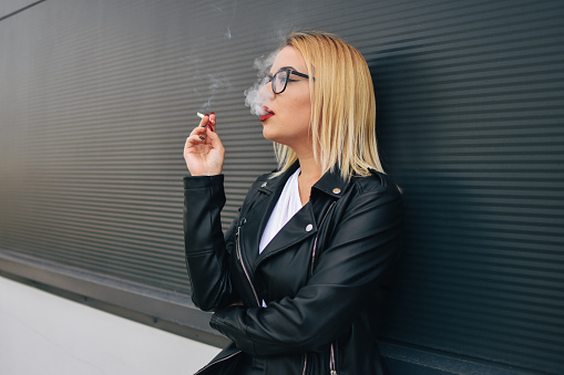 outdoors portrait of a 45 year old blonde woman with curly hair in a black leather jacket and sunglasses smokes a cigarette