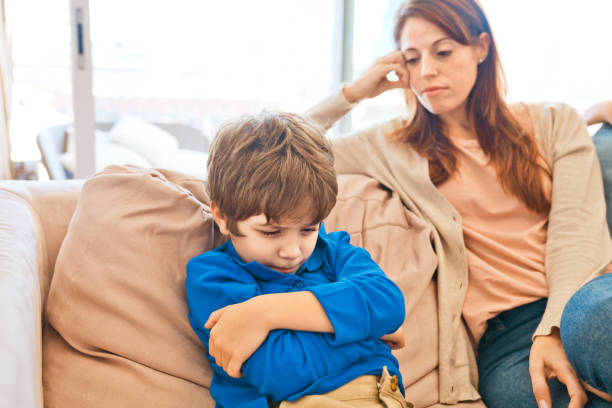 Conflict between mother and son Displeased son sitting with arms crossed on sofa at home. Worried mother looking at her child. Focus on boy. 6 7 years stock pictures, royalty-free photos & images