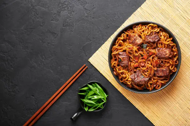 Photo of Ram-Don or Chapaguri noodles with beef steak in black bowl on dark slate background. Jjapaguri is a popular south korean dish with ramen and udon noodles and beef steak. Copy Space