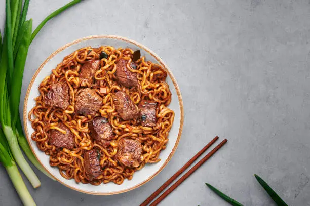 Photo of Ram-Don or Chapaguri noodles with beef steak in white bowl on grey concrete backdrop. Jjapaguri is a popular south korean dish with ramen and udon noodles and beef steak. Copy Space