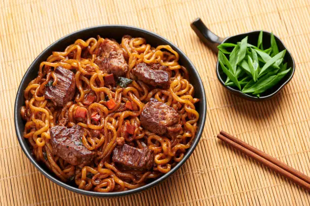 Photo of Ram-Don or Chapaguri noodles with beef steak in black bowl. Jjapaguri is a popular south korean dish with ramen and udon noodles and beef steak.