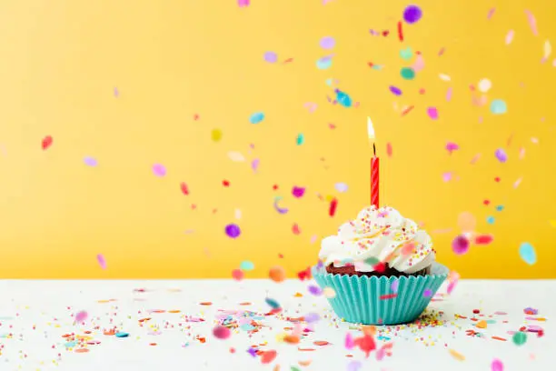 A colorful  birthday cupcake with one candle and confetti on a yellow background