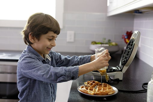A little boy pours honey onto a fresh waffle.  He is five years old and is wearing a long sleeved blue shirt.  They are in a home kitchen with white tiles and white cabinets.  They are preparing the waffle on a black countertop.  He is looking at the honey he is squeezing onto the waffle.  There is a waffle iron next to him.