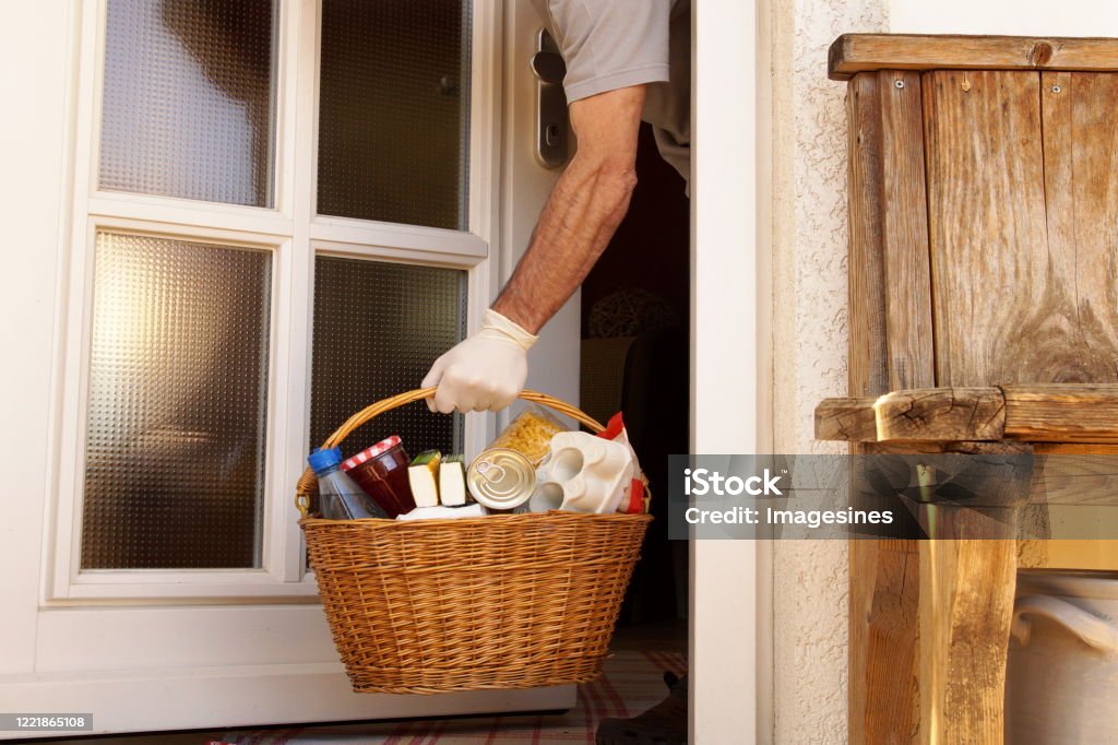 hand of man is taking a full willow wicker basket with handle at he front door. home delivering some groceries at quarantine time because of coronavirus infection COVID-19 Food delivery concept. neighborhood Assistance during Coronavirus Pandemic - Illness Neighbor Stock Photo