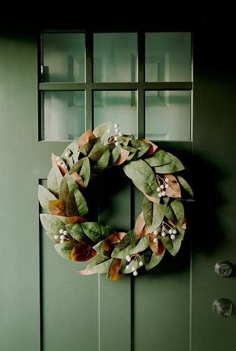 A faux magnolia wreath hangs on a hunter green painted craftsman style front door with front door opened to interior.
