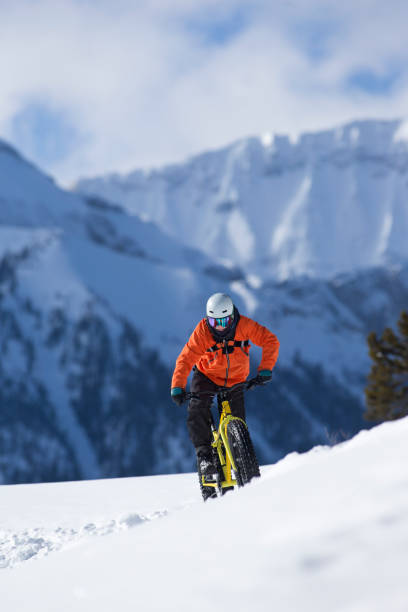 A young man goes for a winter fat bike ride in the Rocky Mountains of Canada. He is wearing warm winter clothing, a downhill ski helmet, and carries a backpack. Fat bikes are mountain bikes with oversized wheels and tires for riding on the snow.
