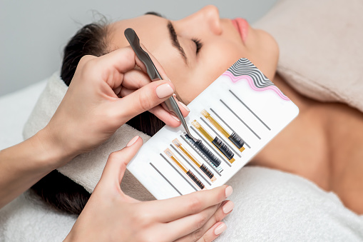 Close up hands of beautician taking artificial eyelashes by tweezers background on face of young woman.