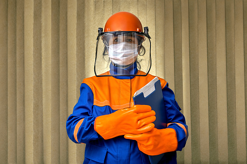 Woman builder uses personal protective equipment and workwear or safety gear during pandemic of coronavirus infection covid-19. Female foreman in a construction helmet, facemask, and face shield.