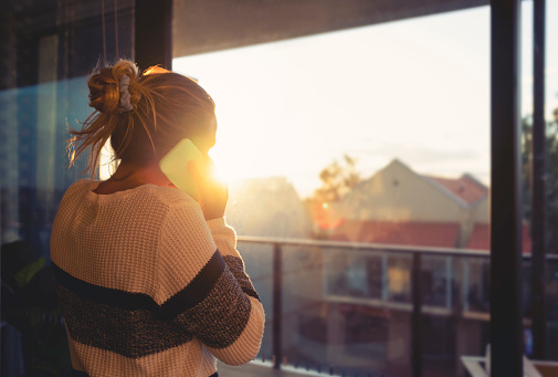 Woman looking through the window at sunset. She is alone and looks a little sad or depressed. She is talking on her mobile phone.