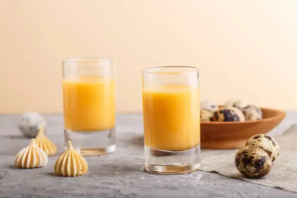Sweet egg liqueur in glass with quail eggs and meringues on a gray and orange background. Side view, close up.