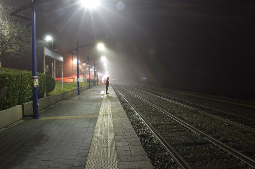 Night view of outdoor train station. Foggy and misty. One man standing on platform, red jacket.