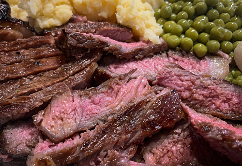 Sliced prime beef steak, cooked medium-rare, with a side of peas and mashed potatoes.