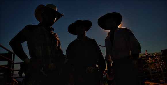Three Men in Silhouette Wearing Cowboy Hats at a Competitive Bull Riding Event at Night