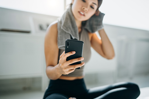 Young Asian woman wiping sweat with a towel around her neck and using smartphone after exercising at home in the morning