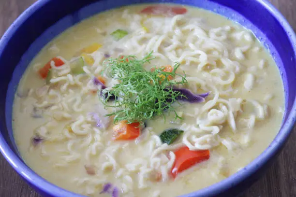 Photo showing a bowl of homemade spicy thai noodle soup / broth isolated on a blue background. This is a low calorie meal made with chopped vegetables and rice noodles, which are low in carbohydrate. This dish is part of a healthy eating, low fat diet plan.