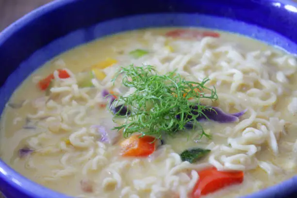 Photo showing a bowl of homemade spicy thai noodle soup / broth isolated on a blue background. This is a low calorie meal made with chopped vegetables and rice noodles, which are low in carbohydrate. This dish is part of a healthy eating, low fat diet plan.