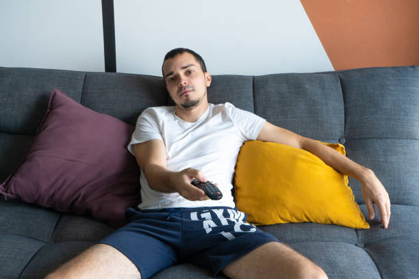 istock A thin guy in home clothes sits on a sofa and watches TV. Bored guy with tv remote control 1221970792