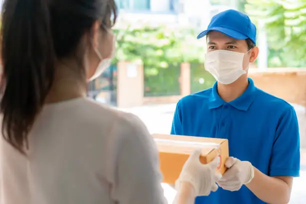 Asian delivery man wearing face mask and glove in blue uniform holding a cardboard boxes in front house and woman accepting a delivery of boxes from deliveryman during COVID-19 outbreak.